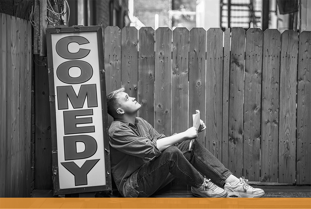 Jess, an adult man living with beck muscular dystrophy, leaning against a comedy sign